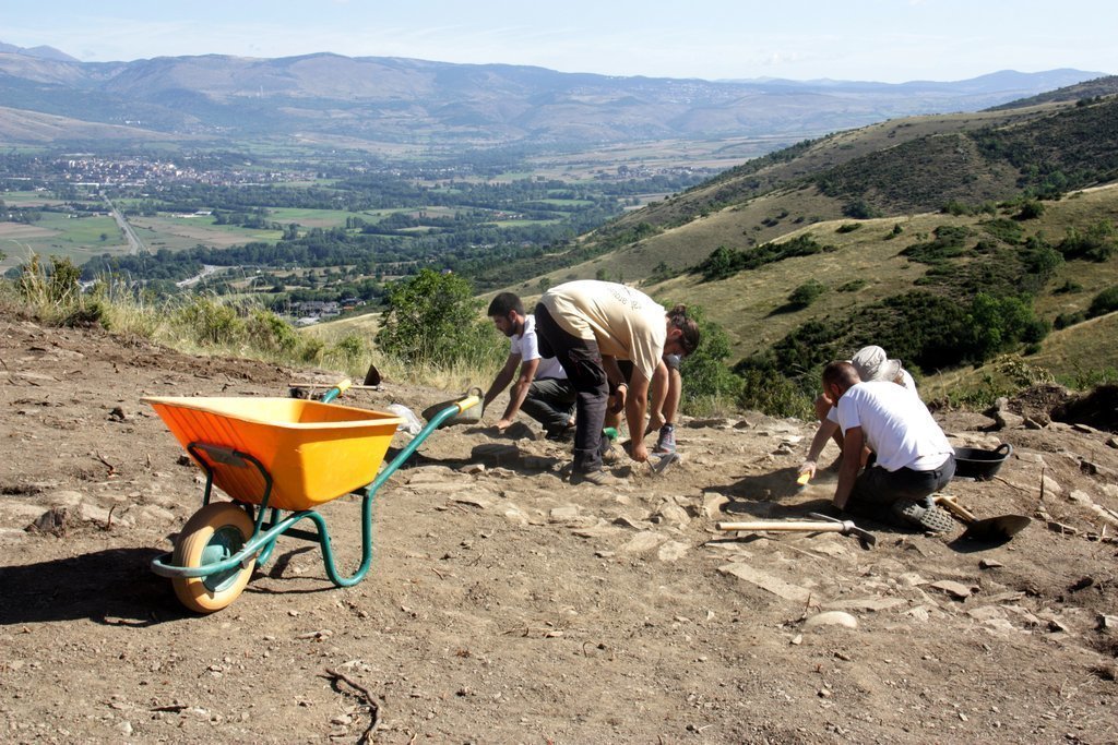 Pla general d'un grup d'arqueòlegs excavant el jaciment del Serrat del Castellar, a Fontanals de Cerdanya, on es veu part de la vall que hi queda a sota del turó. Imatge del 29 d'agost de 2019 (Horitzontal).