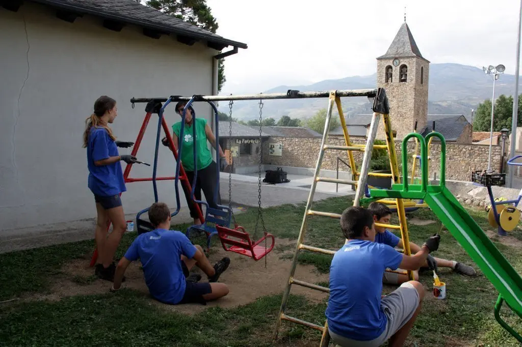 Pla general d'un grup de participants del camp de treball de Bellver de Cerdanya pintant un dels gronxadors del poble de Pi, on es veu l'esgl&radic;&copy;sia al fons, el 27 d'agost de 2019 (Horitzontal).