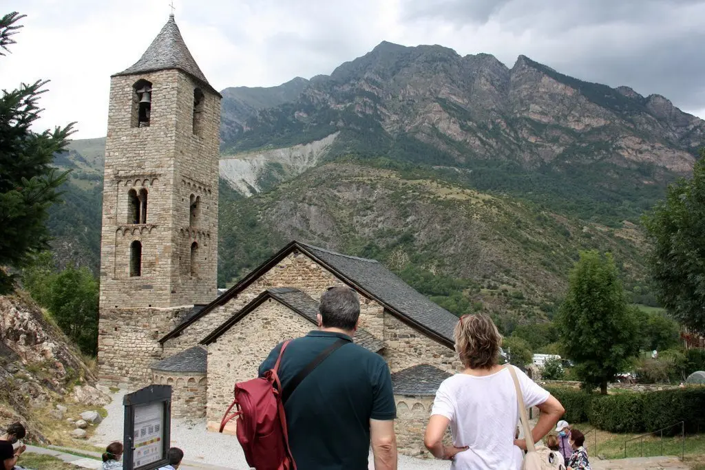 Pla obert de l'esgl&eacute;sia de Sant Joan de Bo&iacute;, a la Vall de Bo&iacute; (Alta Ribagor&ccedil;a), on es veu en primer terme dos turistes accedint-hi al seu entorn i un cartell on hi ha els codis QR per comprar les entrades per Internet, el 14 d'agost de 2020 (Horitzontal).