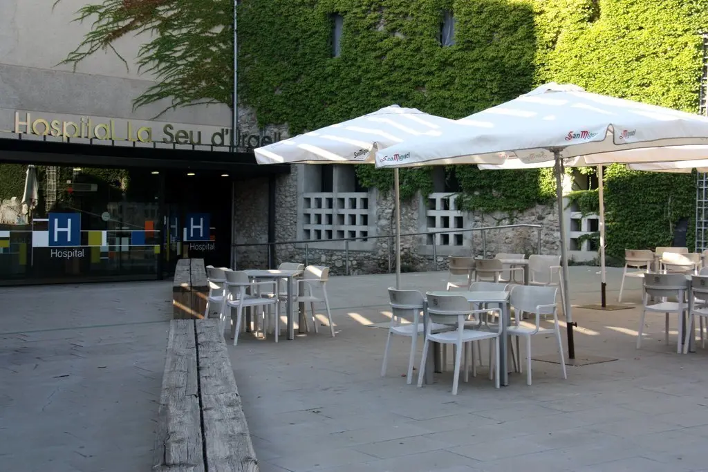 Pla obert on es veu l'exterior del Sant Hospital de la Seu d'Urgell i on hi ha diverses taules d'una terrassa d'un bar col&middot;locades en primer terme. Imatge del 20 d'agost de 2020 (Horitzontal).