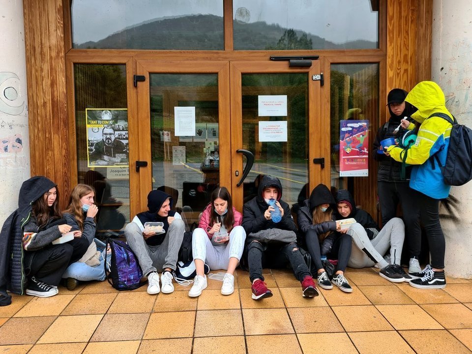 Pla general d'un grup de joves dinant amb carmanyola a la porta de l'Arxiu Comarcal del Pallars Sobirà on es resguardaven de la pluja el 2 d'octubre del 2020. (horitzontal)