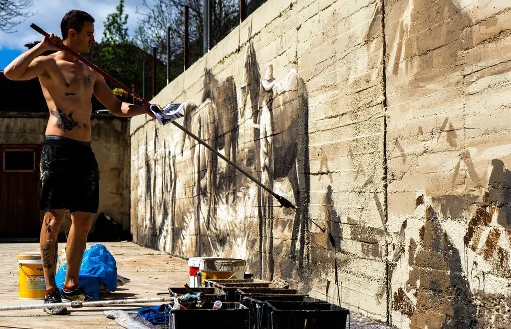 Pla obert de l'artista Nil Safont pintant un mural en una paret de la pista poliesportiva de Tavascan, al Pallars Sobir&agrave;, amb imatges de les feines del camp de fa un segle. Imatge del 19 de maig de 2021. (Horitzontal).