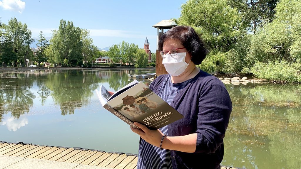 Pla mitjà de l'escriptora Sandra Adam fullejant el seu nou llibre 'L'estiueig a la Cerdanya' on es veu el llac de Puigcerdà al fons. Imatge publicada el 14 de juliol de 2021. (Horitzontal)