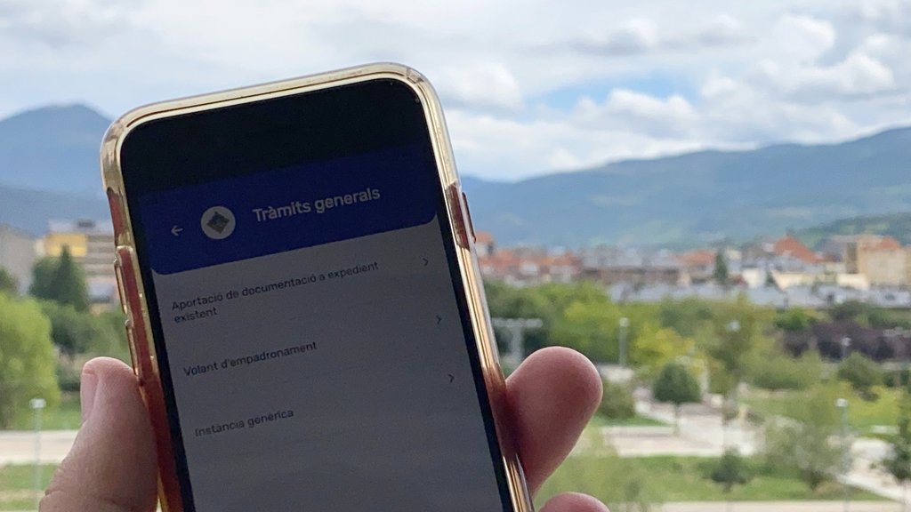 Pla de detall d'una pantalla de l'aplicació de mòbil per fer tràmits municipals a la Seu d'Urgell 'Tramit' on es veu la ciutat al fons. Imatge del 16 de setembre de 2021. (Horitzontal)