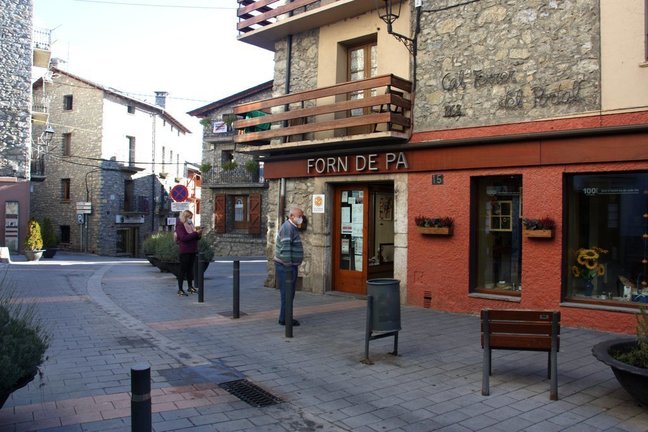 Pla obert d'un carrer de Bellver de Cerdanya on hi ha diversos comerços i on es veu dues persones esperant per entrar al forn de pa en el primer dia de confinament municipal de cap de setmana. Imatge del 30 d'octubre de 2020 (Horitzontal).