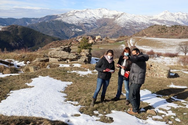 Pla general dels responsables del Museu de Camins mirant els tríptics d'una ruta de senderisme de la Guerra Civil a la Vall de Siarb on es veu darrere els búnquers i el nucli de Vilamur, a Soriguera (Pallars Sobirà), el 20 de gener de 2021 (Horitzontal).