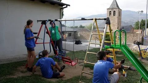 Pla general d'un grup de participants del camp de treball de Bellver de Cerdanya pintant un dels gronxadors del poble de Pi, on es veu l'esgl&radic;&copy;sia al fons, el 27 d'agost de 2019 (Horitzontal).
