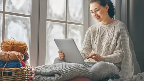 Happy beautiful woman watching videos or enjoying entertainment content on a laptop sitting near windows in the house in the winter.
