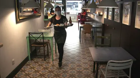 Pla general d'una cambrera d'una cafeteria de la Seu d'Urgell portant una comanda a una de les taules de la terrassa i on es veu que a l'interior del local no n'hi ha cap d'ocupada en aquell moment. Imatge del 25 de maig de 2020 (Horitzontal).