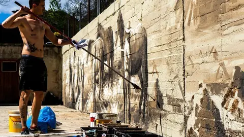 Pla obert de l'artista Nil Safont pintant un mural en una paret de la pista poliesportiva de Tavascan, al Pallars Sobir&agrave;, amb imatges de les feines del camp de fa un segle. Imatge del 19 de maig de 2021. (Horitzontal).