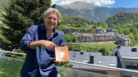 Pla mitj&agrave; de l'escriptor Greg Coonen mostrant la portada del seu llibre 'La P&iacute;fia' i on es veu part del nucli andorr&agrave; de La Massana i el paisatge natural que l'envolta al fons. Imatge del 2 de juny de 2021. (Horitzontal)