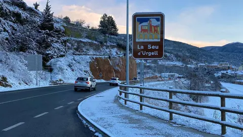 Pla obert de diversos vehicles circulant per la zona d'entrada a la Seu d'Urgell per l'N-260, on es veu un cartell indicatiu de la ciutat, la cal&ccedil;ada totalment neta i el paisatge emblanquinat despr&eacute;s de la nevada, el 29 de novembre de 2021. (Horitzontal)