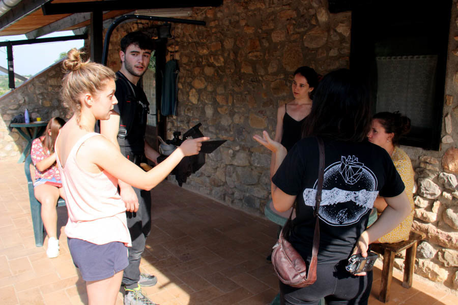 Pla americà on es veuen alguns dels participants del Laboratori de Creació Audiovisual del Cerdanya Film Festival comentant l'escena que gravaran del seu curtmetratge fora d'una casa de colònies de Bellver de Cerdanya, el 7 d'agost de 2019 (Horitzontal).