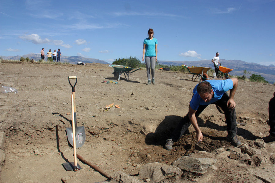 Pla general d'un arqueòleg excavant el jaciment del Serrat del Castellar, a Fontanals de Cerdanya, on es veuen diverses persones del mateix equip al seu darrere. Imatge del 29 d'agost de 2019 (Horitzontal).