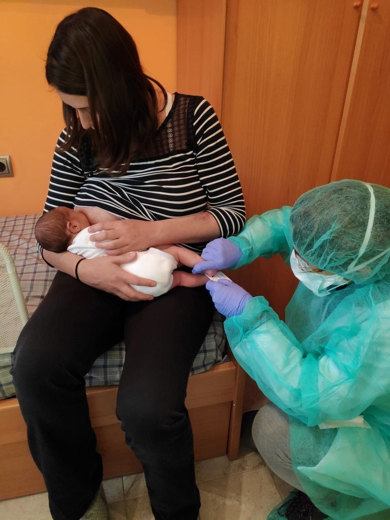 Pla general d'un nadó amb la seva mare i una professional de l'equip de Pediatria dels Pirineus en una visita a domicili. Imatge facilitada per Pediatria dels Pirineus el 12 d'abril de 2020 (Vertical).