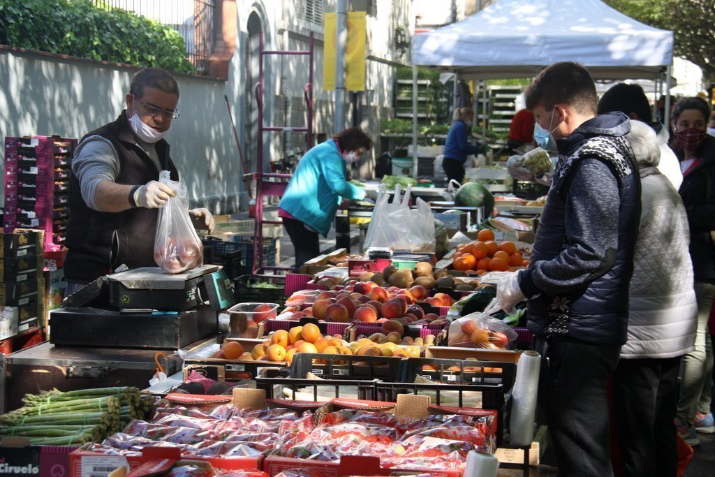 Pla americà d'un paradista de fruites i verdures del mercat de la Seu d'Urgell mentre despatxa a un client complint amb les mesures de seguretat per evitar la propagació de la covid-19. Imatge del 28 d'abril de 2020 (Horitzontal).
