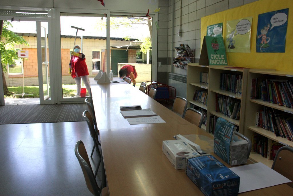 Pla general de tots els equips de protecció damunt de les taules que es faran servir a l'escola Alfons I de Puigcerdà per al tràmit de la preinscripció i dues persones netejant els vidres de la sala al darrere. Imatge del 18 de maig de 2020 (Horitzontal).