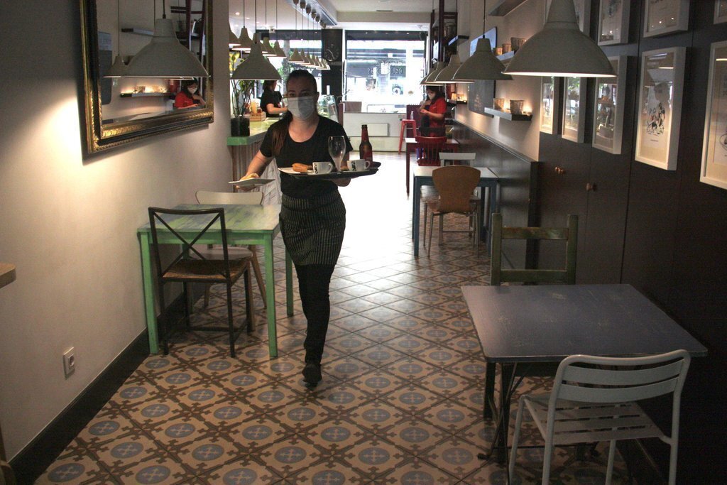 Pla general d'una cambrera d'una cafeteria de la Seu d'Urgell portant una comanda a una de les taules de la terrassa i on es veu que a l'interior del local no n'hi ha cap d'ocupada en aquell moment. Imatge del 25 de maig de 2020 (Horitzontal).