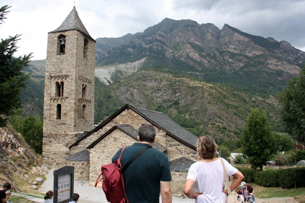 Pla obert de l'església de Sant Joan de Boí, a la Vall de Boí (Alta Ribagorça), on es veu en primer terme dos turistes accedint-hi al seu entorn i un cartell on hi ha els codis QR per comprar les entrades per Internet, el 14 d'agost de 2020 (Horitzontal).