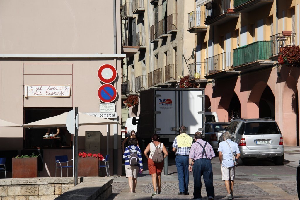 Pla obert on es veuen diverses persones d'esquena caminant pel carrer Major de la Seu d'Urgell i una taula d'una terrassa de bar al fons. Imatge del 20 d'agost de 2020 (Horitzontal).