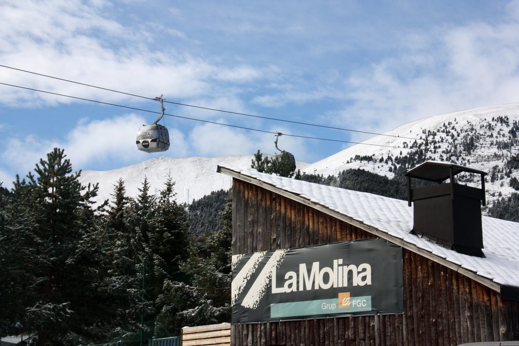 Pla obert d'un cartell indicatiu de l'estació de La Molina col·locat a l'exterior d'un edifici i on es veuen al fons la muntanya nevada i dos cabines del telecabina. Imatge del 15 d'octubre de 2020 (Horitzontal).