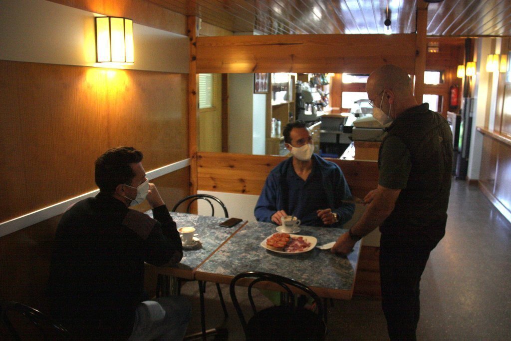 Pla americà de dos clients asseguts en una taula d'un bar del nucli d'Adrall, a Ribera d'Urgellet (Alt Urgell), on es veu un dels propietaris de l'establiment servint-los menjar. Imatge del 23 de novembre de 2020 (Horitzontal).