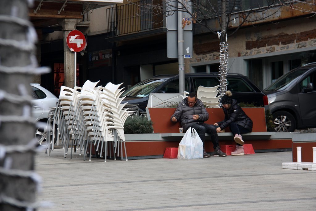 Pla obert on es veuen dues persones assegudes en un banc de la plaça Cabrinetty de Puigcerdà amb un cafè per emportar en el primer dia de confinament perimetral de la Cerdanya. Imatge del 23 de desembre de 2020 (Horitzontal).