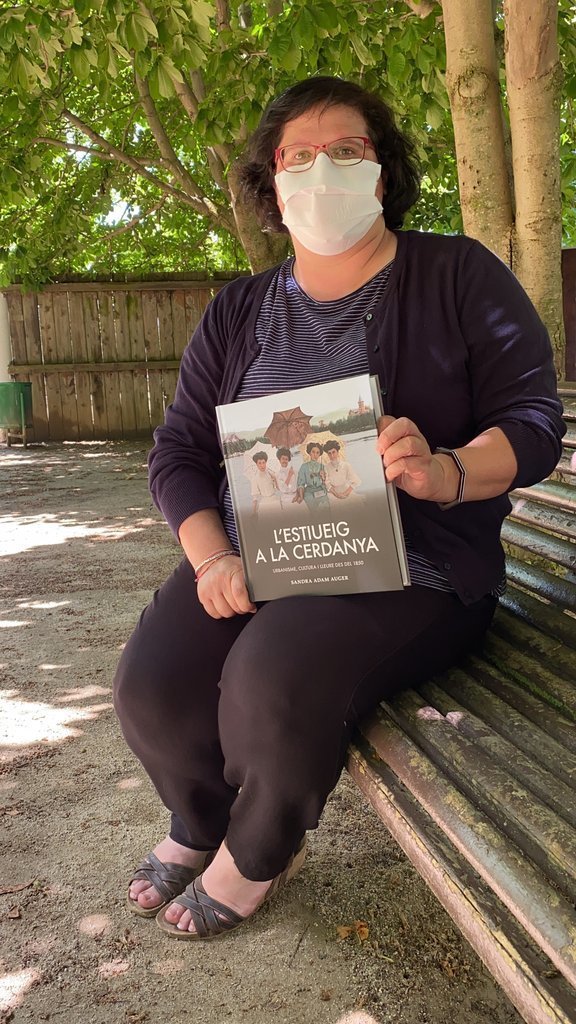 Pla general de l'escriptora Sandra Adam asseguda en un banc del Parc Schierbeck de Puigcerdà mostrant el seu nou llibre 'L'estiueig a la Cerdanya'. Imatge publicada el 14 de juliol de 2021. (Vertical)