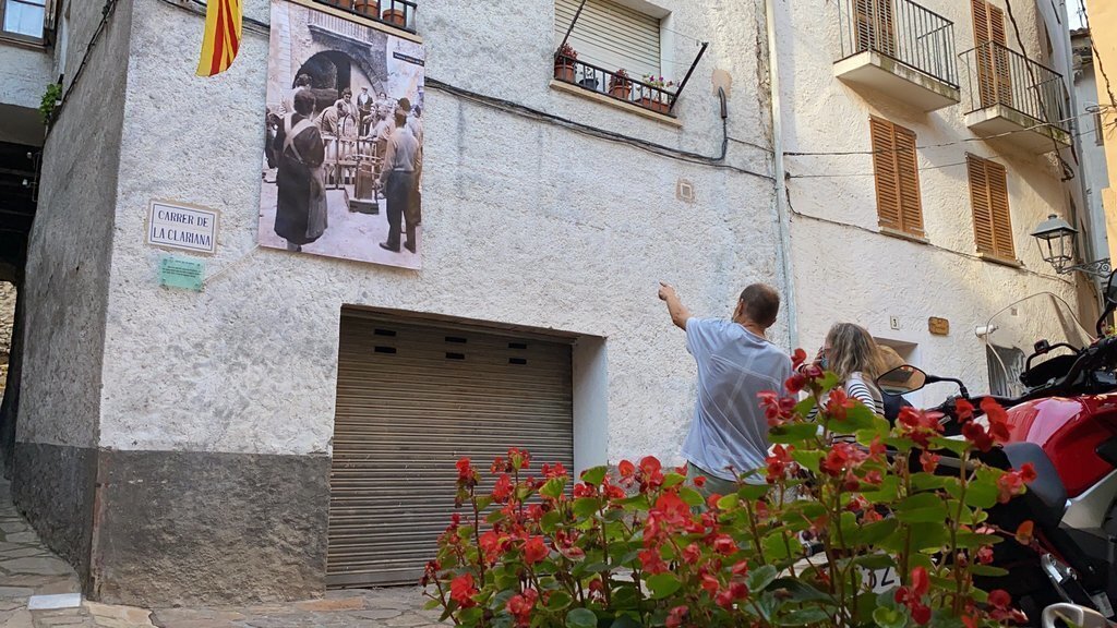 Pla obert on es veu una pancarta amb una fotografia antiga col·locada en un dels carrers de Peramola (Alt Urgell) que forma part de la ruta de memòria històrica i on es veuen diverses persones observant-la. Imatge del 19 d'agost de 2021. (Horitzontal)