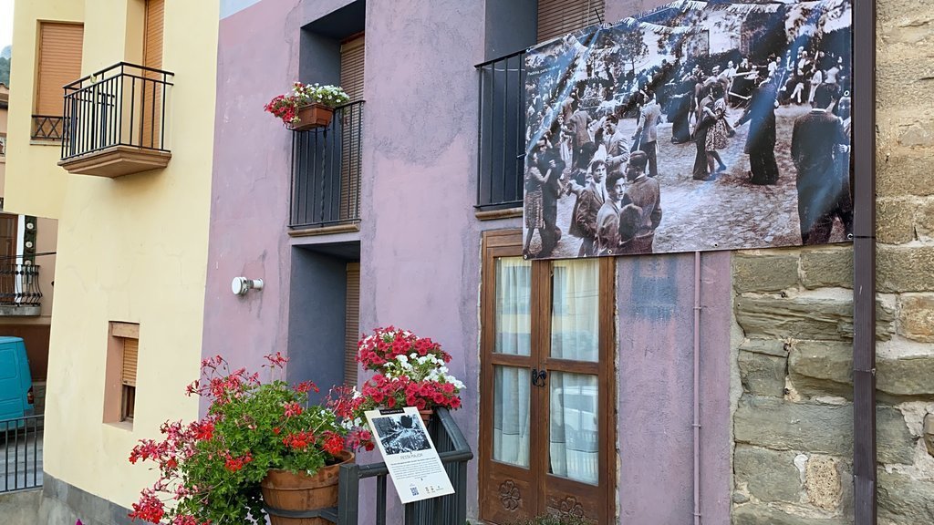 Pla obert on es veu una pancarta amb una fotografia antiga col·locada en una plaça de Peramola (Alt Urgell) i la placa informativa d'un dels punts de la ruta de memòria històrica. Imatge del 19 d'agost de 2021. (Horitzontal)