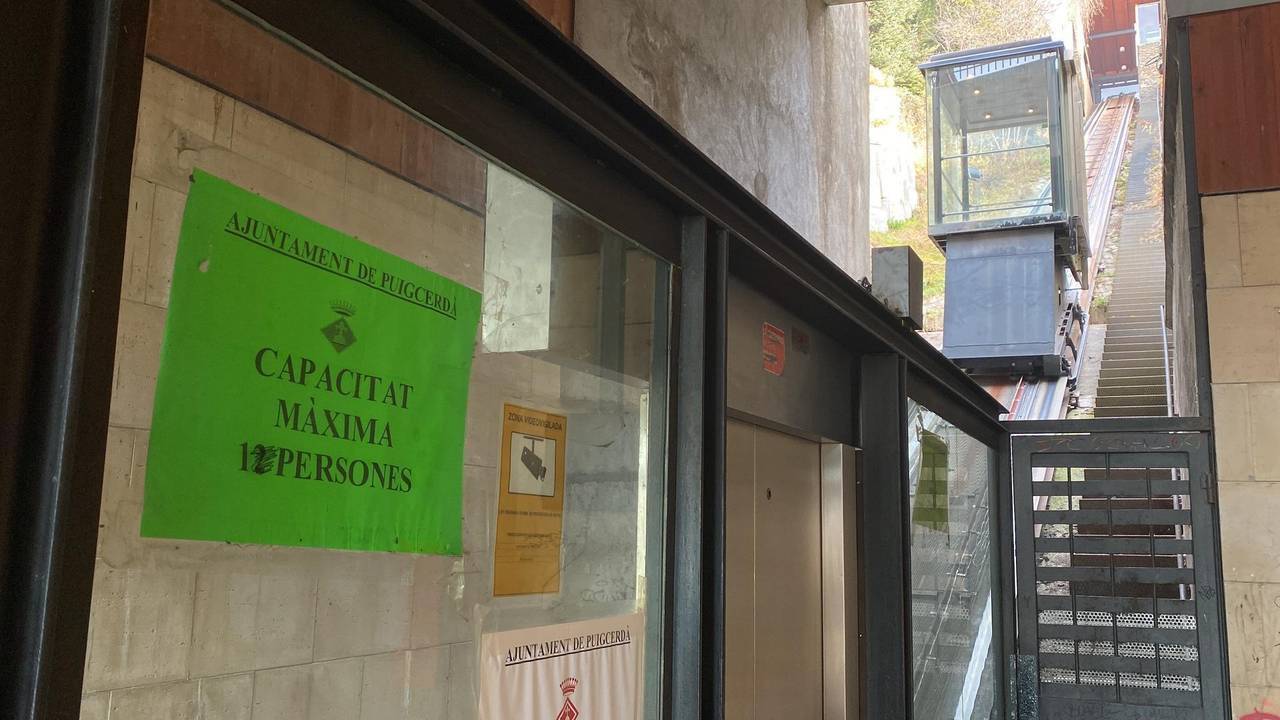 Pla de detall de l'ascensor inclinat de Puigcerdà arribant a la zona de l'estació de tren on es veu part de les portes d'accés i un cartell que indica la seva capacitat màxima. Imatge del 4 de gener de 2022. (Horitzontal).